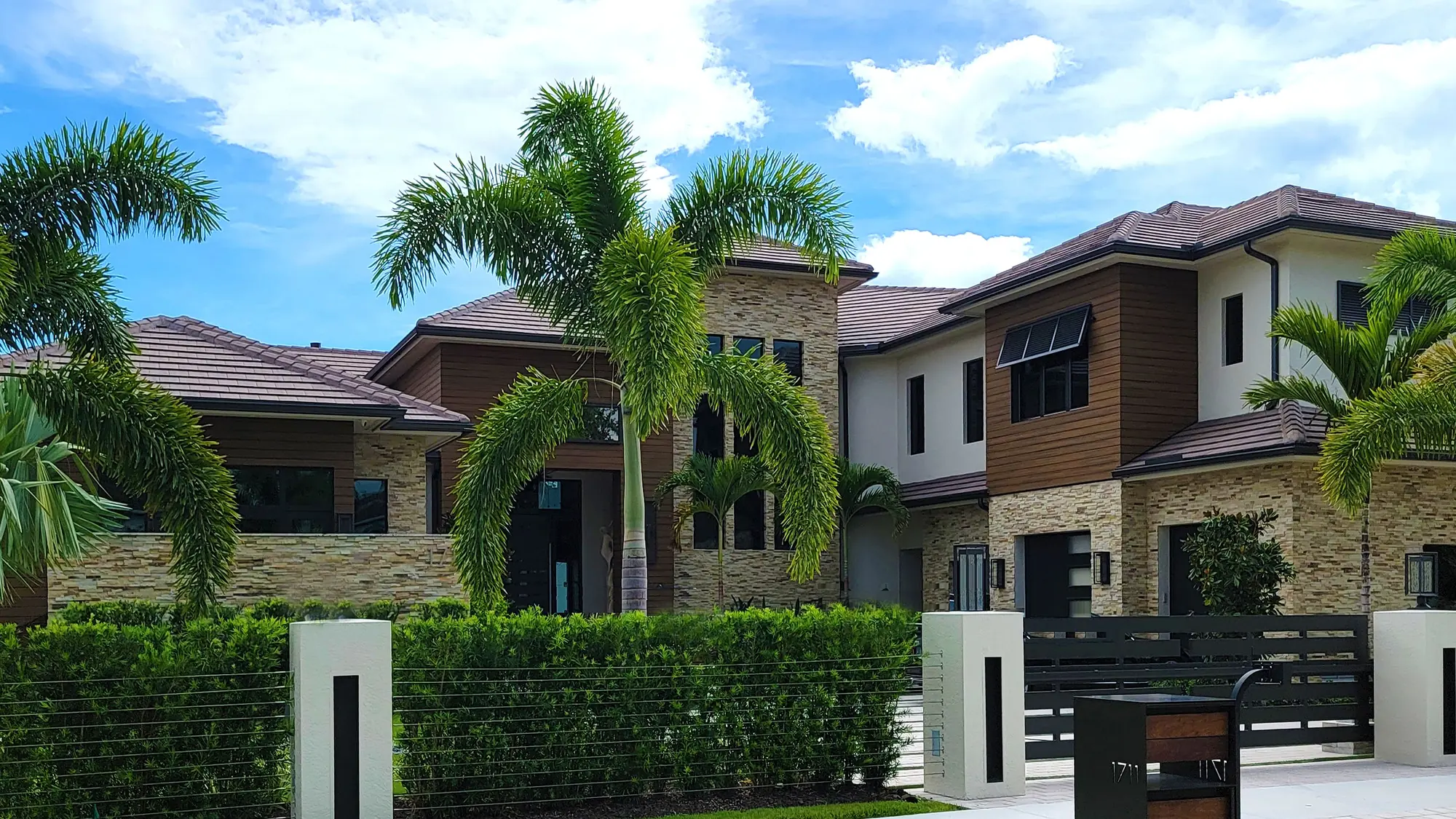 A large modern waterfront home in Florida, with walls of glass, wood cladding and tall palms along the seawall.