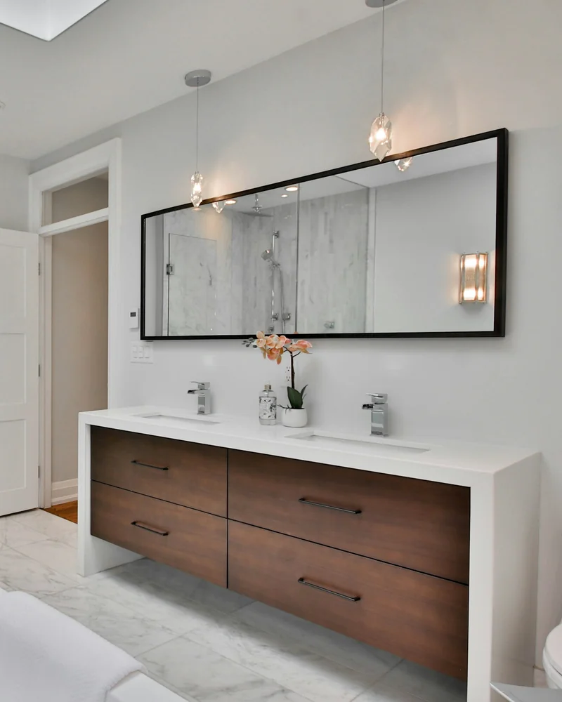 A modern bathroom with a floating walnut double vanity, a stone counter and a marble tiled walk in shower reflected in the mirror.
