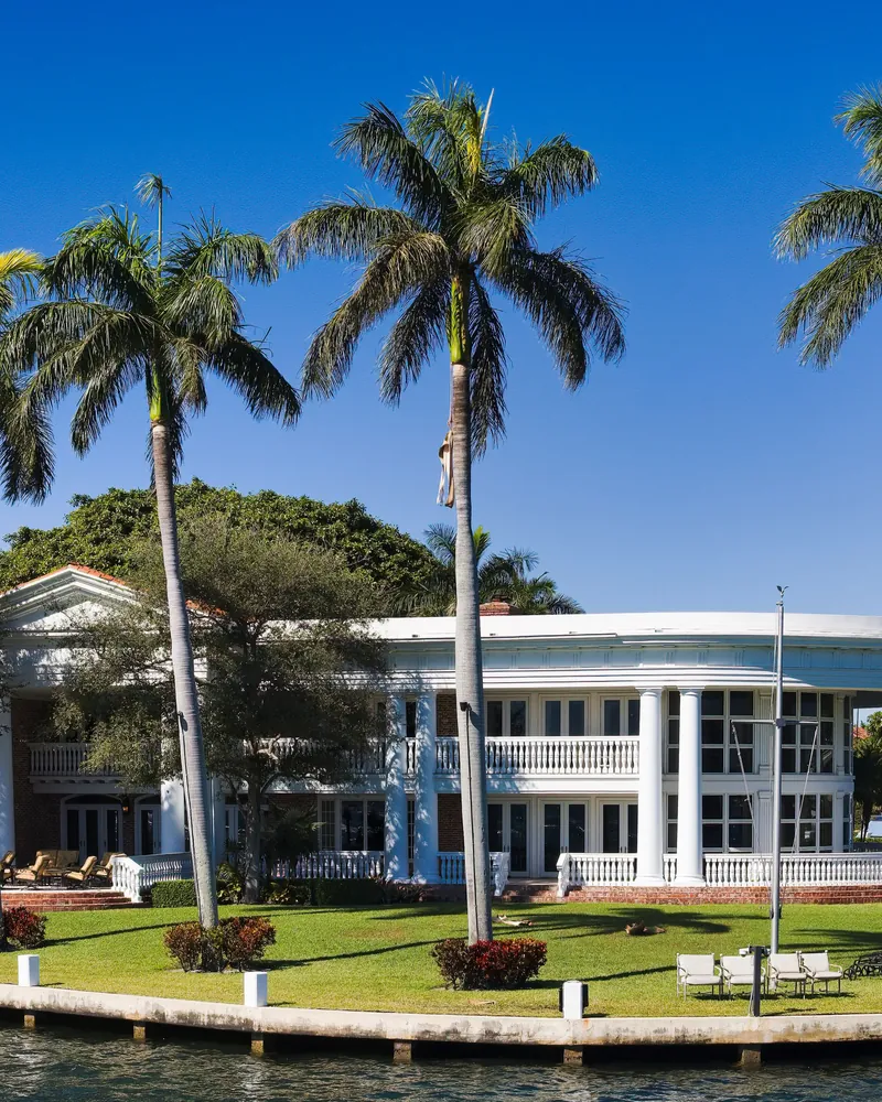 A large white waterfront home behind a lawn and tall royal palms, seen from the water.