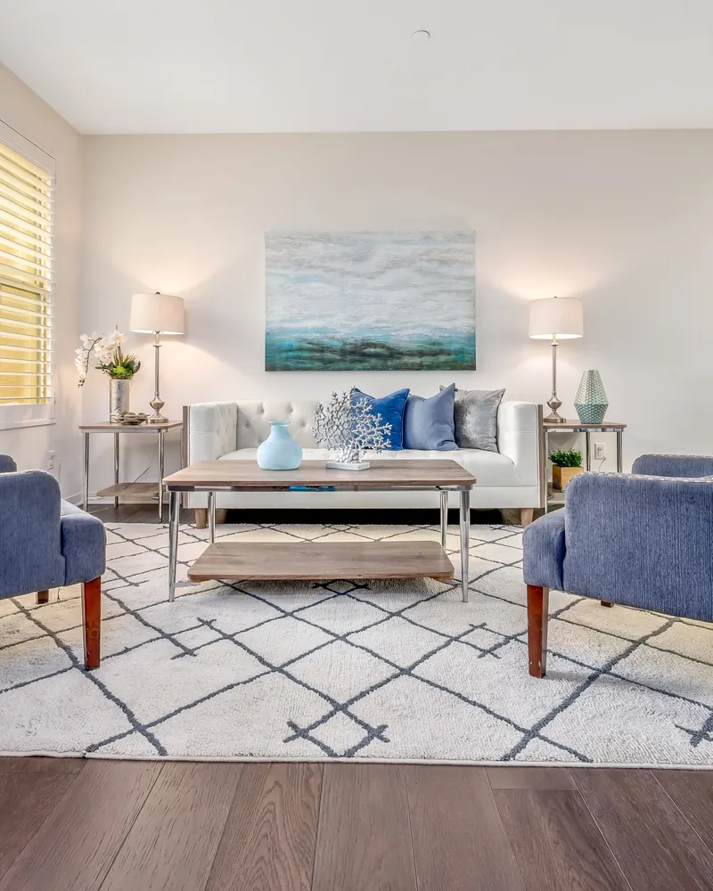 A bright coastal living room styled for listing photographs, with blue armchairs and a seascape hung above the sofa.